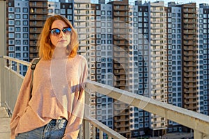 Tween redhead girl in pullover, jeans and sunglasses standing on balcony against high-rise multi-storey residential building at