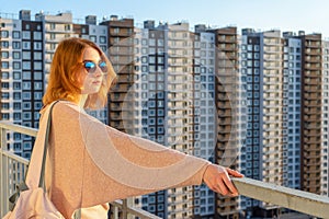 Tween redhead girl in pullover, jeans and sunglasses standing on balcony against high-rise multi-storey residential building at