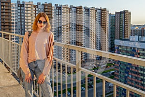 Tween redhead girl in pullover, jeans and sunglasses standing on balcony against high-rise multi-storey residential building at