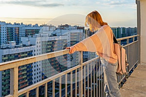 Tween redhead girl in pullover, jeans and sunglasses standing on balcony against high-rise multi-storey residential building at