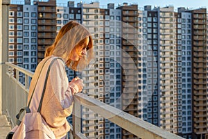 Tween redhead girl in pullover, jeans and sunglasses standing on balcony against high-rise multi-storey residential building at