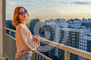 Tween redhead girl in pullover, jeans and sunglasses standing on balcony against high-rise multi-storey residential building at