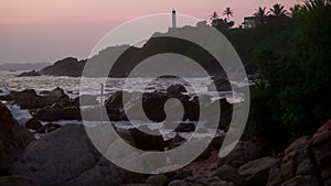 Twilight descends on rocky coastline sea waves crash over boulders near lighthouse. Palm trees sway against pinkish sky