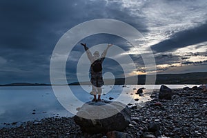 Twilight. A full-length man stands on a rock near the water, greeting the sunset