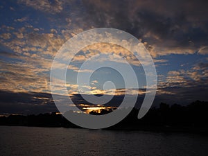 Twilight landscape of sun sets over the forest on a riverbank, blue sky covered with dark thunder clouds