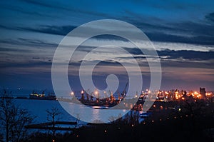 Twilight over industrial harbor with cargo cranes, lit buildings, operational port area. Dusk silhouette skyline