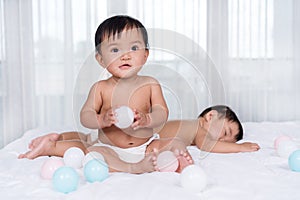 Twin babies on bed, one playing color ball, one sleeping