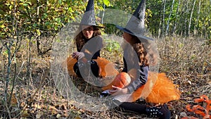Twin girls dressed like witches are seating in a forest and laughing.
