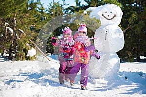 Twin girls in the winter in the woods for a walk near a large snowman. Children in pink jackets and glasses in the sun jumping