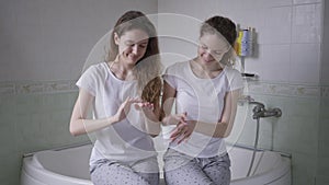 Twin sisters in pajamas sitting on bathtub applying moisturizer on hands smiling. Medium shot portrait two confident