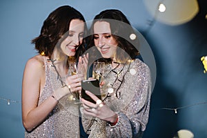 Twin sisters in shiny evening dresses celebrate with glasses of sparkling wine