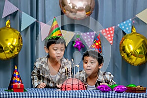 Twins adorable boy in shirt, celebrating his birthday, blowing candles on homemade baked cake, indoor. Birthday party for kids