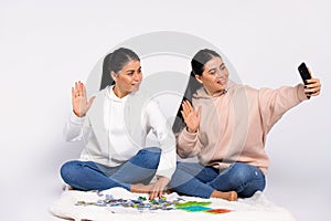 Twins are sitting on the floor, putting together a puzzle and communicating via video call on a smartphone. White