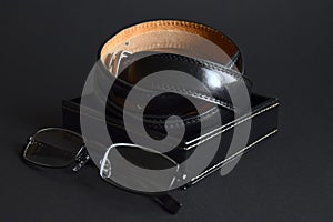 Twisted men`s leather belt in black in a box and eyeglasses on dark background, elegant accessories