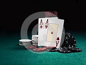 Two aces hearts and diamonds standing leaning on chips piles, some of them laying nearby on green cover of playing table