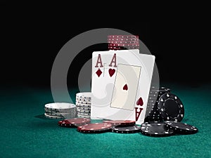 Two aces hearts and diamonds standing leaning on chips piles, some of them laying nearby on green cover of playing table