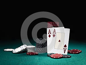 Two aces hearts and diamonds standing leaning on chips piles, some of them laying nearby on green cover of playing table