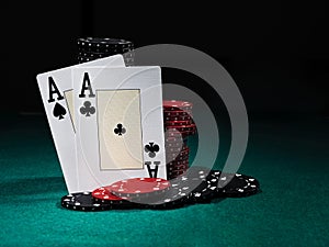 Two aces standing leaning on chips piles, some of them laying nearby on green cover of playing table. Black background