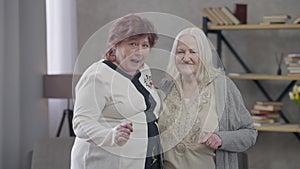 Two active senior women dancing in living room looking at camera. Portrait of positive female retirees having fun