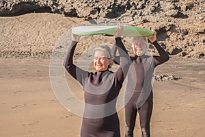 Two active seniors holding a surfboard and wearing wetsuits to go surf - mature people or pensioners trying to learn surfing