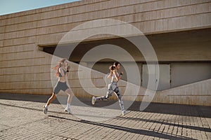 Two active women athlete running on sunny morning side by side on modern buildings background