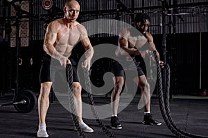 two active young men with battle rope doing exercise in functional training fitness gym.