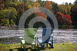 Two Adirondack chairs sitting on a river shore with trees in the background