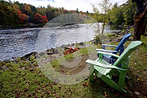 Two Adirondack chairs sitting on a river shore with trees in the background