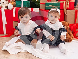 Two adorable babies sitting on floor by christmas gifts at home