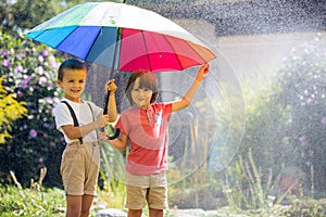 Two adorable children, boy brothers, playing with colorful umbrella under sprinkling water