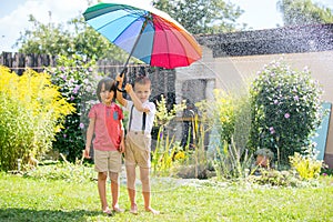 Two adorable children, boy brothers, playing with colorful umbrella under sprinkling water