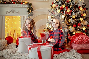 Two adorable girls in checkered dresses sitting near Christmas tree and opening their presents