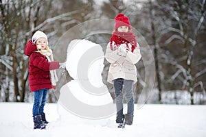 Two adorable little girls building a snowman together in beautiful winter park. Cute sisters playing in a snow.