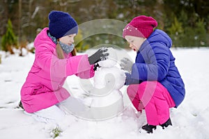 Two adorable little girls building a snowman together in beautiful winter park. Cute sisters playing in a snow.