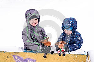 two adorable preschool kids brother boys in winter wear sit amoung snow and play with toy reindeer