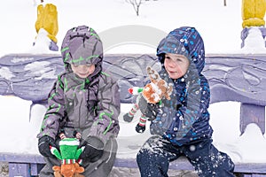 two adorable preschool kids brother boys in winter wear sit amoung snow and play with toy reindeer