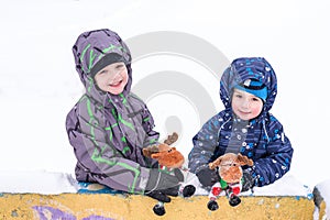two adorable preschool kids brother boys in winter wear sit amoung snow and play with toy reindeer