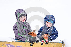 two adorable preschool kids brother boys in winter wear sit amoung snow and play with toy reindeer