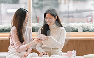 Two adorable young long hair women wearing sweater, chatting together, holding mobile phone, smiling with happiness, sitting in