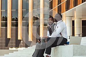 Two African American businessmen in suits communicate. Friends and business partners sit on the concrete steps of the