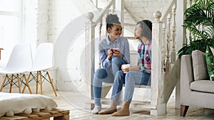 Two african american curly girls sistres sitting on stairs have fun laughing and chatting together at home