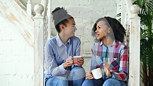 Two african american curly girls sistres sitting on stairs have fun laughing and chatting together at home