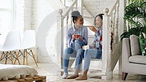 Two african american curly girls sistres sitting on stairs have fun laughing and chatting together at home
