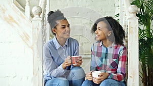 Two african american curly girls sistres sitting on stairs have fun laughing and chatting together at home