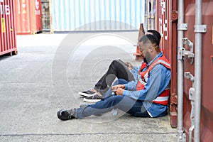 Two African American logistic staff workers wearing safety vests sitting on the floor against the container take a break after