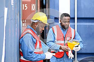 Two African American logistic staff workers wearing safety vests stand against the container and take a break after hard work.
