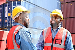 Two African American logistic workers wearing reflective vests and white helmets talk about logistics operations at shipping