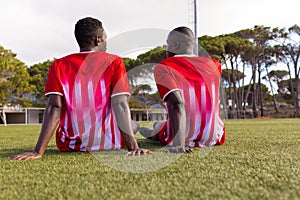 Two African American young male athletes are sitting on grass on field outdoors, wearing red jerseys