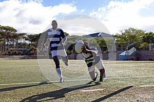 Two African American young male athletes in striped jerseys play rugby under the sun on a field