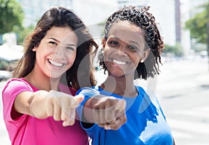 Two african and caucasian women pointing at camera in the city